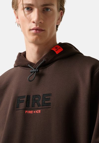 Młody mężczyzna w brązowej bluzie z kapturem, z haftem "FIRE" w kolorze czarnym i "FIRE + ICE" w kolorze czerwonym poniżej, z czerwonym metką na kapturze.