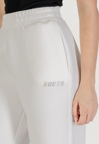 Pantaloni jogger bianchi in cotone con vita elasticizzata, tasche laterali e logo "GUESS" in strass argentati sulla coscia sinistra.