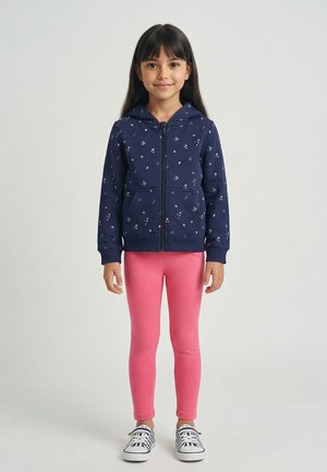 Jong meisje staand, gekleed in een marineblauwe zip-up hoodie met bloemenprint, felroze legging en witte sneakers met marineblauwe strepen.