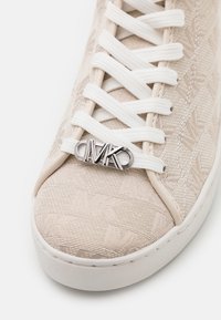 Sneaker beige bermotif tekstru, dengan kain berpola, tali putih, dan aksen logo perak di lidah sepatu. Sol karet dengan finishing yang halus.