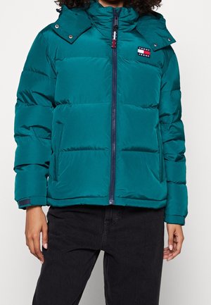 Teal puffer jacket with front zipper, side pockets, hood, and Tommy Jeans logo patch on left chest, worn with black pants.