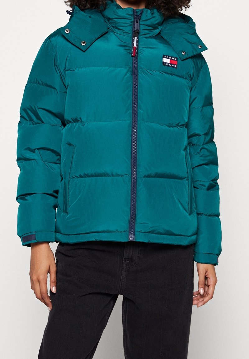 Doudoune bleu sarcelle avec fermeture éclair à l'avant, poches latérales, capuche et patch logo Tommy Jeans sur la poitrine gauche, portée avec un pantalon noir.