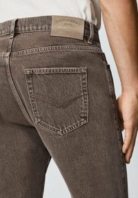 Jeans in denim marrone con una tasca posteriore standard caratterizzata da dettagli di cucitura sottili e un'etichetta in pelle beige sulla vita.