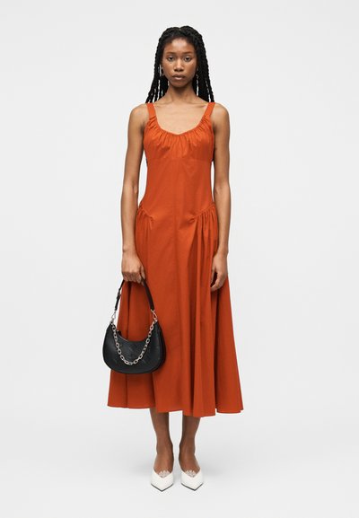 Γυναίκα που φοράει μίντι φόρεμα χωρίς μανίκια σε σκουρό πορτοκαλί χρώμα, κρατώντας μια μαύρη τσάντα με ασημένια αλυσίδα και φορώντας λευκές μυτερές γόβες.