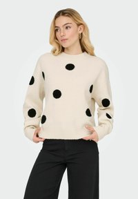 Pull en maille crème avec grands pois noirs, encolure ronde et ourlet côtelé. Le modèle le porte avec un pantalon noir sur un fond clair.