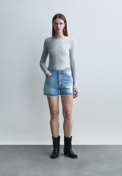 Νεαρή γυναίκα που στέκεται με το ένα χέρι στην τσέπη, φορώντας ανοιχτό γκρι μπλούζα με μακριά μανίκια, σορτς από τζιν και μαύρα μποτάκια μέχρι τον αστράγαλο σε απλό φόντο.