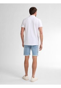 Camisa polo blanca, pantalones cortos de mezclilla azul claro remangados en los puños y zapatos color crema. Detalles de costura visibles en la camisa y en los bolsillos de los pantalones cortos.