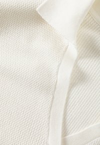 Tissu texturé de couleur crème avec un motif gaufré et des accents lisses. Il présente un col côtelé et des bords doux et tissés.
