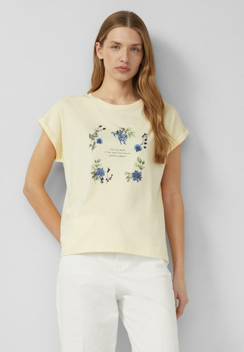 Frau mit langen hellbraunen Haaren trägt ein blassgelbes T-Shirt mit blauem Blumenmuster und weißen Hosen, steht vor einem schlichten grauen Hintergrund.