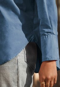 Camicia blu a maniche lunghe con orlo curvo, realizzata in tessuto morbido. Presenta maniche arrotolate e vestibilità casual, abbinata a denim grigio chiaro.