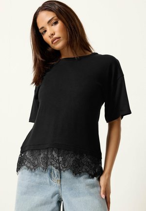 Vrouw draagt een zwarte top met korte mouwen met kanten zoom en lichtblauwe jeans, poseert tegen een effen lichte achtergrond.