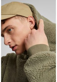 Olivgrüne Fleecejacke mit hohem Kragen und strukturierter Oberfläche; nackte Haut sichtbar im Gesicht, mit einer beigefarbenen Baseballmütze.
