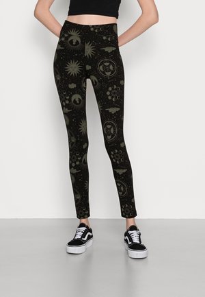 Zwarte leggings met groene sterrenbeelden, met zonnen, wolken en geometrische vormen. Model draagt zwarte sneakers.