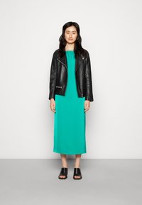 Jaqueta de motociclista de couro preta sobre um vestido teal fluído, com uma silhueta reta e uma bainha até ao chão. Sandálias deslizadas pretas completam o visual.