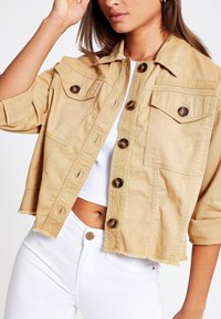 Vaaleanbeige farkkutakki, jossa on lyhyt malli, terävä kaulus, kaksi rintataskua ja suuret ruskeat napit. Refrattuja helmoja.