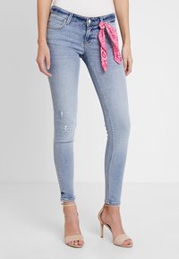 Jeans ajustados de mezclilla en un tono azul claro con un diseño desgastado, con un pañuelo de estampado rosa atado en la cintura. Llevados con sandalias de tacón en color nude.