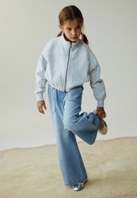 Sivi dukser sa zatvaračem, svijetloplave široke hlače s džepovima i srebrne baletanke, stoji na mekanom bež površini.