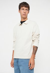 Cămașă polo bej deschis cu mânecă lungă, cu guler clasic și trei nasturi, asortată cu blugi din denim bleu deschis, având un tatuaj pe antebraț.