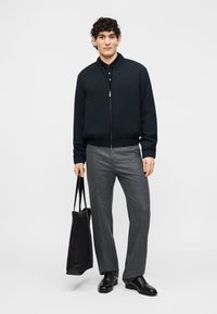 Jeune homme portant une veste noire à fermeture éclair, un pantalon gris, des chaussures noires, tenant un sac fourre-tout noir sur un fond blanc uni.