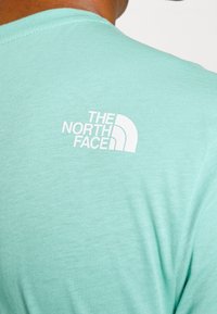 Miętowa koszulka z krótkim rękawem wykonana z miękkiego materiału, z białym logo w kształcie półokręgu na górnej części pleców.