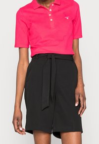 Růžová polo tričko s krátkým rukávem a třemi knoflíky, spárované s černými šortkami s vysokým pasem, které mají uvázaný pásek a volný střih.