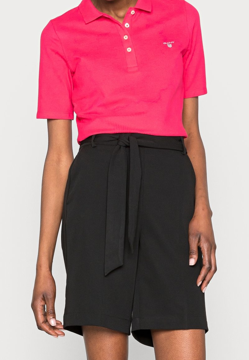 Růžová polo tričko s krátkým rukávem a třemi knoflíky, spárované s černými šortkami s vysokým pasem, které mají uvázaný pásek a volný střih.