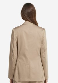 Beige blazer med en åtsittande design, slät yta, axelsömmar och två bakventiler. Har en notch-krage och långa ärmar.