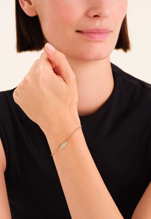 Pulsera de oro con un colgante alargado y liso, incrustado con una piedra preciosa brillante, asegurada con una delicada cadena. Se lleva en la muñeca.