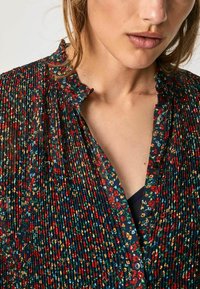 Blusa floreale con una texture plissettata. Presenta uno sfondo blu navy, piccoli fiori rossi e gialli, e un colletto arricciato con chiusura a bottoni.