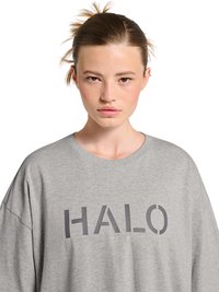 Młoda osoba z zaczesanymi do tyłu włosami, nosząca szarą, oversize koszulę z dużym napisem "HALO" na przodzie.