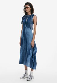 Blue textured dress with a keyhole detail, asymmetrical hem, paired with metallic sneakers and white ankle socks. Accented with a blue wrap.