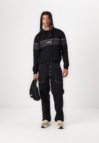 Sudadera negra con detalles grises y logotipo, combinada con pantalones negros con costuras blancas. El modelo sostiene un bolso negro pequeño y lleva zapatillas.