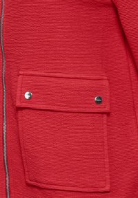 Giacca rossa a trama con una grande tasca frontale dotata di bottone a scatto e una zip argento a contrasto. Il materiale presenta un motivo ondulato.