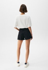 T-shirt bianco oversize, pantaloni shorts in denim nero a vita alta con risvolti, sneakers bianche con dettagli blu navy; sfondo neutro.