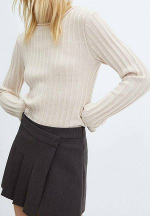 Kvinde iført en beige ribstrikket langærmet sweater, stoppet ned i en mørkegrå plisseret miniskørt, med hænderne hvilende på hoften.