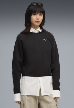 Sort cropped sweatshirt med ribbet tekstur, prydet med hvidt logo. Kombineret med en hvid skjorte med knapper, hvor kraven og manchetterne er synlige.