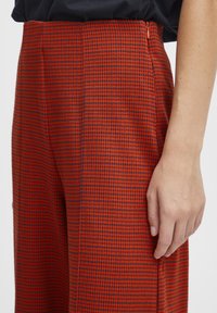 Pantalons à motif pied-de-coq rouge et noir avec une texture lisse, dotés d'une fermeture éclair sur le côté et d'une coupe décontractée, présentés au niveau de la taille.