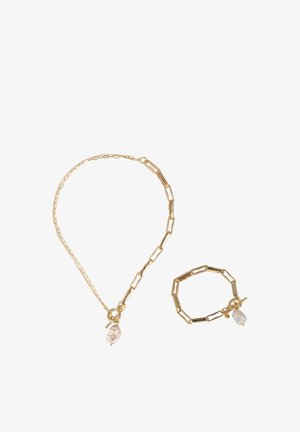 Ensemble collier et bracelet en chaîne dorée, avec des maillons allongés et un accent en pierre transparente sur chaque pièce. Texture lisse et design solide.