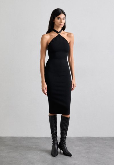 Schwarzes Neckholderkleid mit taillierter Silhouette und knielangem Saum aus strukturiertem Material. Kombiniert mit knielangen schwarzen Stiefeln mit Absatz.