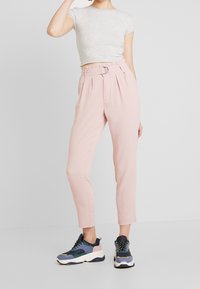 Pantalons tailleur rose clair, avec une taille ceinturée, des plis et des jambes fuselées, associés à des baskets chunky multicolores et un haut gris ajusté.