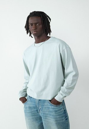 Himmelblaues Langarmshirt aus Baumwolle, mit lockerer Passform, gerippten Bündchen und kombiniert mit hellblauen, lässig vernieteten Jeans.