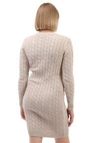 Vrouw met kort blond haar die een beige kabelbreisel lange mouw trui-jurk draagt, met de rug naar ons gekeerd tegen een witte achtergrond.