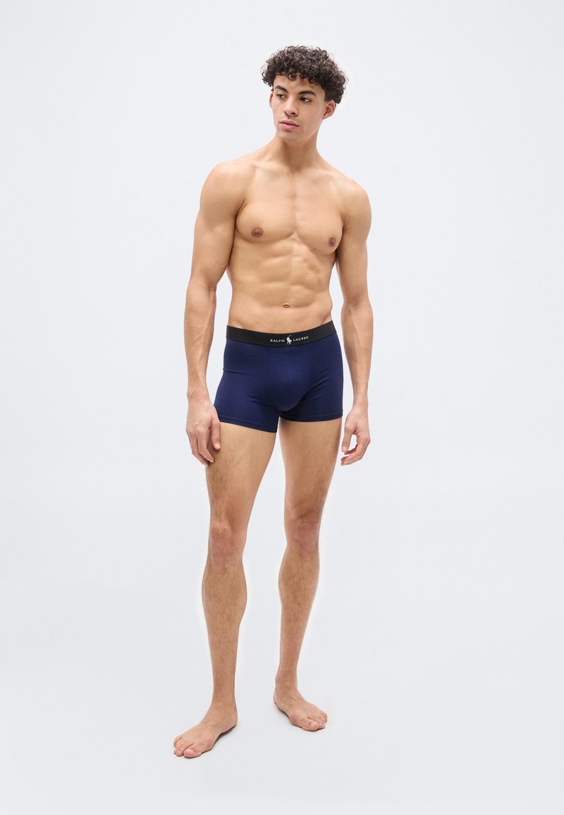 Νέος άνδρας όρθιος, ξυπόλητος, φοράει σκούρο μπλε εσώρουχα boxer με μαύρη μέση ζώνη, κοιτάζει προς τα πλάγια σε απλό φωτεινό φόντο.