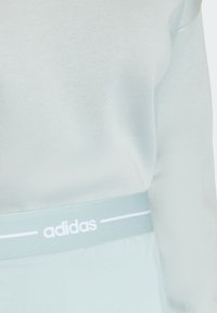 Svetlomodrá mikina so hladkou textúrou má kontrastný biely pás s výrazným logom Adidas.