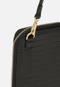Sac bandoulière noir avec motif crocodile embossé, présentant une finition texturée, une fermeture éclair dorée et une attache à clip sur la bandoulière en cuir.