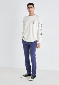 Cămașă crema cu mânecă lungă și imprimeuri grafice negre, asociată cu pantaloni albaștri și adidași din pânză negri cu talpă albă.