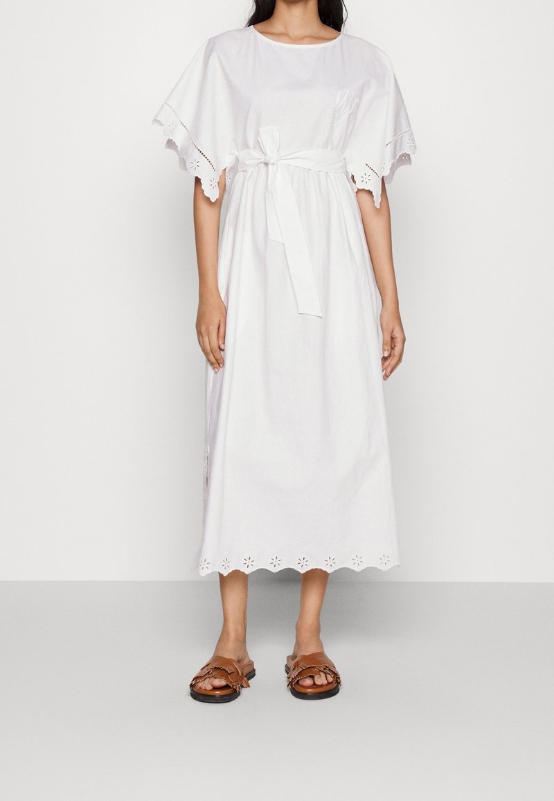 Femme portant une robe midi blanche avec des bords festonnés, une ceinture nouée à la taille et des manches papillon, associée à des sandales marron.