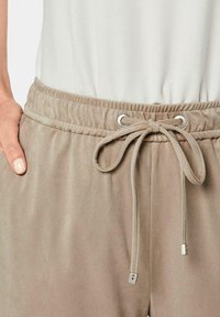 Beige trainingsbroek met een elastische tailleband en een trekkoord. De stof lijkt zacht met een gladde textuur, voorzien van zilverkleurige oogjes en aglets.