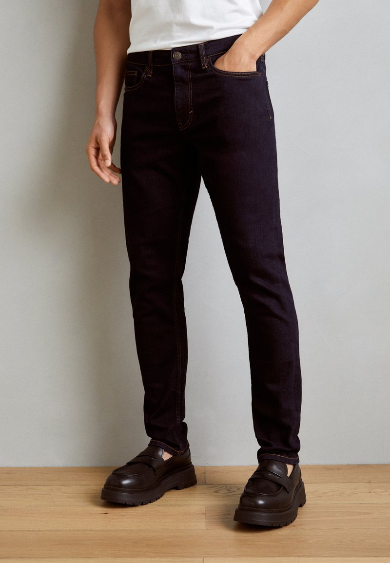 Jeans de mezclilla azul oscuro con un ajuste slim, que presentan costuras naranjas en contraste y un cierre de botón, combinados con zapatos negros de suela gruesa.