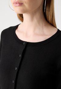 Nainen, joka on pukeutunut mustaan nappimalliseen neulepuseroon, jossa on pyöreä kaula-aukko, korvalehdissä renkaat ja suorat vaaleanruskeat hiukset, yksinkertaista taustaa vasten.